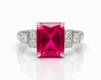 May include: A silver ring with a large, rectangular, bright pink gemstone at its centre. The ring is accented with smaller, clear gemstones on either side. The design is set against a white background, creating a striking contrast and elegant look.