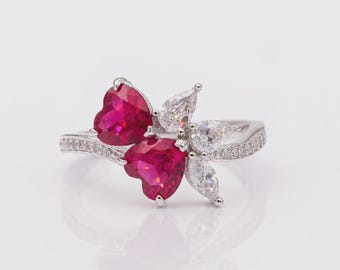 May include: A silver-toned ring featuring two heart-shaped vibrant red stones and three clear pear-shaped stones arranged in a cluster. The band is slender and encrusted with small clear stones along the visible portion of the metal shank.