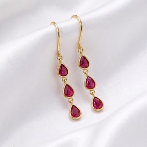 May include: A pair of gold drop earrings, each featuring three teardrop-shaped red gemstones suspended in a vertical line. The earrings are attached to simple gold hooks, showcasing a polished metal finish and vibrant red stones.
