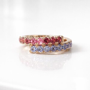 May include: A gold ring featuring two overlapping bands, one set with a row of round pink and red gemstones and the other set with a row of round light blue gemstones, creating a layered appearance with a polished metallic finish.