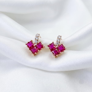 May include: A pair of gold-tone stud earrings, each featuring three square-cut vibrant pink stones arranged in a heart-like cluster, accented by a single small clear stone on the post, creating a symmetrical and polished jewellery design.