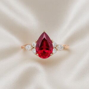 May include: A ring featuring a large, pear-shaped, deep red gemstone set in a gold band. The central stone is flanked by three smaller, clear, marquise-cut diamonds on each side, secured by gold prongs, creating a symmetrical cluster design.