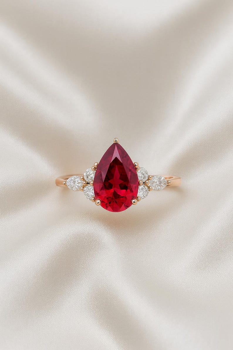 May include: A ring featuring a large, pear-shaped, deep red gemstone set in a gold band. The central stone is flanked by three smaller, clear, marquise-cut diamonds on each side, secured by gold prongs, creating a symmetrical cluster design.