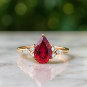 May include: A gold ring featuring a large pear-shaped red gemstone as the centrepiece, flanked by three small round and pear-shaped clear stones on each side. The band is polished gold, and the setting holds the stones securely in place.