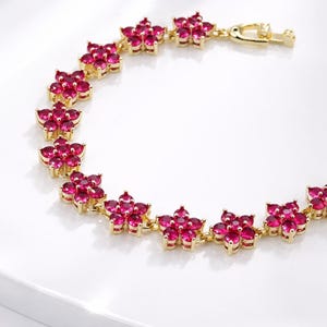 Peut inclure: Un bracelet doré orné de fleurs. Chaque fleur est composée de plusieurs petites pierres précieuses roses vives. Le bracelet est fermé par un fermoir. Le bijou est présenté sur une surface blanche.