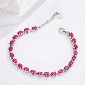 May include: A silver-toned bracelet featuring a series of oval-cut vibrant pink stones set in individual metal frames. The bracelet includes a delicate chain extension with a small heart-shaped charm at the end and a secure lobster claw clasp.