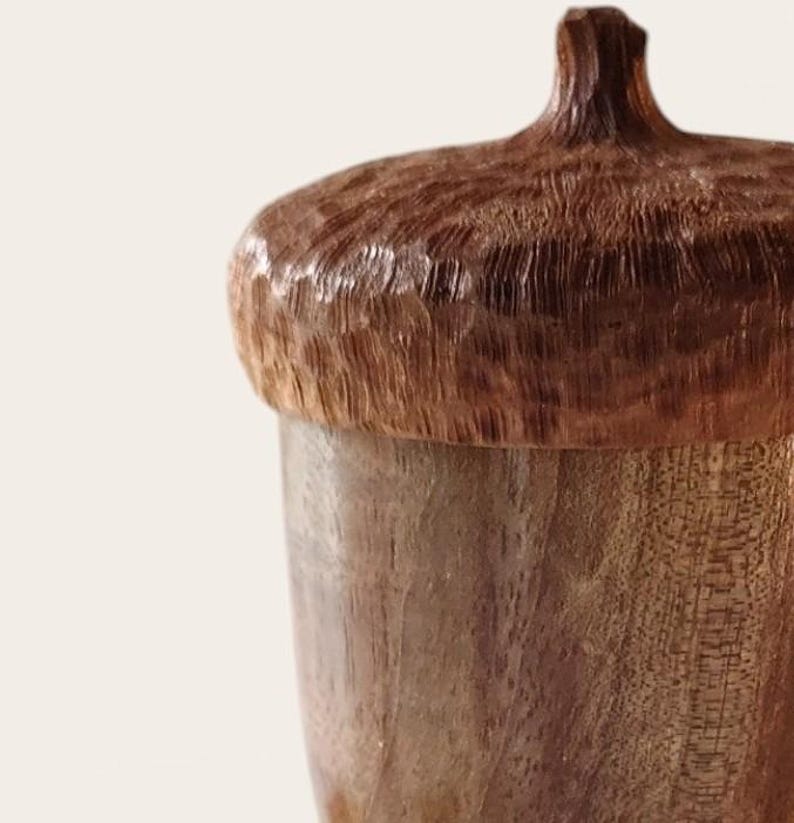 Peut inclure: Un r&eacute;cipient en bois en forme de gland, dot&eacute; d'un capuchon textur&eacute; brun fonc&eacute; avec une petite tige sur le dessus et d'un corps lisse brun plus clair avec des motifs de grain de bois visibles. L'objet est sculpt&eacute; dans du bois naturel.