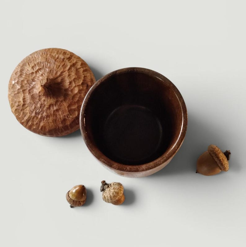 Peut inclure: Un r&eacute;cipient en bois brun fonc&eacute; avec un couvercle amovible pr&eacute;sentant une surface sculpt&eacute;e et textur&eacute;e rappelant un chapeau de gland. Le r&eacute;cipient est ouvert, r&eacute;v&eacute;lant un int&eacute;rieur lisse, et est accompagn&eacute; de trois petits glands naturels.