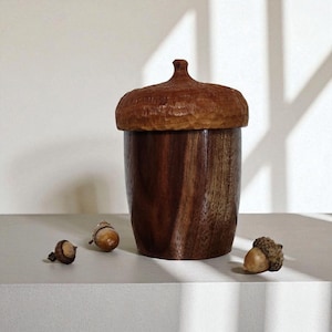 Peut inclure: Un r&eacute;cipient en bois en forme de gland avec un corps brun fonc&eacute; au grain lisse et un chapeau sculpt&eacute; textur&eacute;. Le r&eacute;cipient est accompagn&eacute; de trois petits glands naturels plac&eacute;s autour de sa base, pr&eacute;sentant un design botanique r&eacute;aliste.
