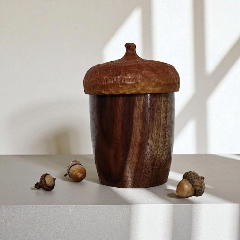 Peut inclure: Un r&eacute;cipient en bois en forme de gland avec un corps brun fonc&eacute; au grain lisse et un chapeau sculpt&eacute; textur&eacute;. Le r&eacute;cipient est accompagn&eacute; de trois petits glands naturels plac&eacute;s autour de sa base, pr&eacute;sentant un design botanique r&eacute;aliste.
