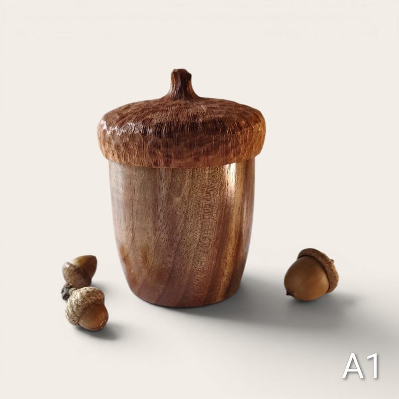 Peut inclure: Un r&eacute;cipient en bois en forme de gland avec un chapeau textur&eacute; brun fonc&eacute; et un corps lisse brun clair. Le r&eacute;cipient est accompagn&eacute; de deux petits glands naturels plac&eacute;s sur la surface. Le texte A1 est visible dans le coin inf&eacute;rieur droit.
