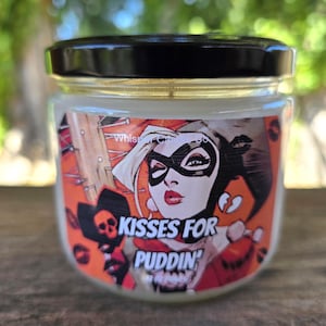 May include: A glass jar candle with a black lid. The label features a comic book style illustration of a woman with a black mask and red lipstick. The text on the label reads "Kisses for Puddin'" in white letters.