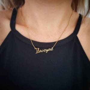 Puede incluir: Un collar dorado con las palabras "I love you" en fuente cursiva. El collar se lleva sobre una parte superior negra, creando un contraste que resalta la joya. La cadena es delicada y el colgante es de estilo script.