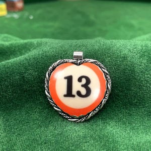 Puede incluir: Un colgante en forma de corazón con un centro de color crema que muestra el número 13 en negro, rodeado por un borde naranja y un marco de metal plateado trenzado. El colgante incluye una pequeña anilla de metal plateado en la parte superior.