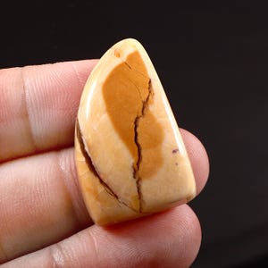 Op de afbeelding: Een gepolijste, druppelvormige edelsteen met een crème- en bruinpatroon. De steen heeft een grote, onregelmatige bruine vlek en een prominente scheur die door het midden loopt. De edelsteen wordt in een hand gehouden.