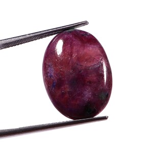 Pode incluir: Uma pedra preciosa polida, de formato oval, com um tom vermelho profundo, acentuada por inclusões roxas e azuis mais escuras. A gema é segurada por uma pinça prateada sobre um fundo branco. A superfície da pedra é lisa e refletora.