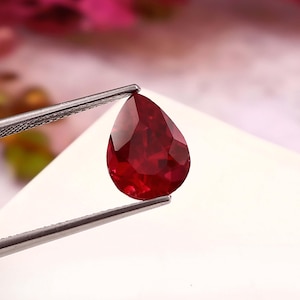May include: A faceted, teardrop-shaped ruby gemstone, held by tweezers. The ruby is a deep, saturated red color with a high degree of clarity and brilliance. The gemstone is set against a white background, highlighting its color and shape.