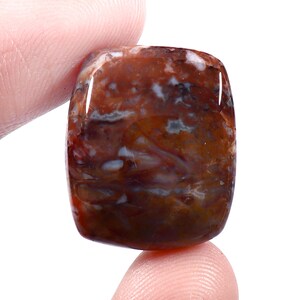 Op de afbeelding: Een gepolijste, rechthoekige edelsteen met een mix van bruine, rode en witte patronen. De steen heeft een glanzend oppervlak en afgeronde randen. De edelsteen wordt tussen twee vingers gehouden.