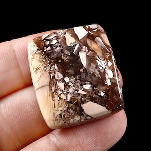 Op de afbeelding: Een rechthoekige edelsteen met een mix van bruin, beige en witte kleuren. De steen heeft een uniek patroon met een mozaïekachtig uiterlijk, met onregelmatige vormen en texturen. De edelsteen wordt in een hand tegen een zwarte achtergrond gehouden.