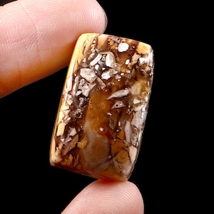 Op de afbeelding: Een rechthoekige edelsteen met een gepolijst oppervlak. De steen heeft een mix van bruine, beige en witte kleuren, met een patroon van onregelmatige vormen en texturen. De edelsteen wordt tussen twee vingers tegen een zwarte achtergrond gehouden.