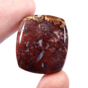 Op de afbeelding: Een gepolijste, vierkante edelsteen met patronen in dieprood, bruin en wit. Tussen vingers gehouden, onthult het een glad, glanzend oppervlak en natuurlijke kleurvariaties. Geschikt voor sieraden of als decoratief item.