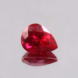 May include: A faceted, pear-shaped ruby gemstone. The gem is a deep, vibrant red colour, with multiple facets catching the light. The ruby is set against a reflective surface, creating a mirrored effect. The overall shape is teardrop-like.