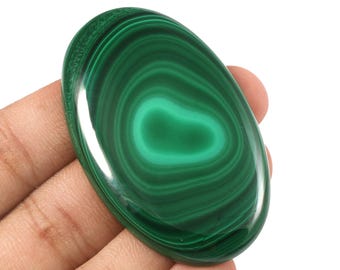 May include: Oval-shaped malachite gemstone with concentric bands of dark and light green. The polished stone is held in a hand, showcasing its natural patterns and vibrant color. The gemstone has a smooth, glossy surface.