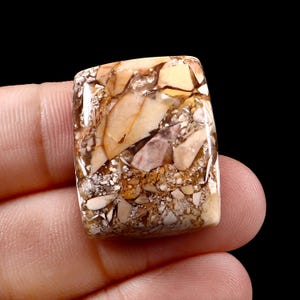 Op de afbeelding: Een rechthoekige edelsteen met een mozaïkpatroon van beige, bruine en crèmekleuren. De steen heeft een gepolijst, glanzend oppervlak en het patroon lijkt op een natuurlijke rotsformatie. De edelsteen wordt in een hand tegen een zwarte achtergrond gehouden.