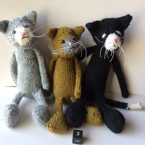 May include: Three knitted cat toys, one gray, one brown, and one black, with white faces and whiskers. The toys are sitting on a white surface with a black number 3 block.