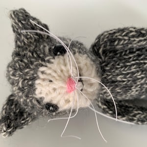 May include: A handmade knitted cat toy in shades of gray and white. The cat has black button eyes, a pink nose, and white whiskers. The toy is lying down, with its head and body visible.