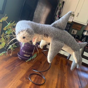 May include: A handmade knitted shark toy in shades of gray and white, with a pink mouth and black eyes. The shark is displayed on a decorative metal stand. The toy is placed on a wooden table.