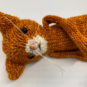 May include: A hand-knitted orange cat toy with white whiskers and black eyes. The cat is lying on its back with its paws in the air.