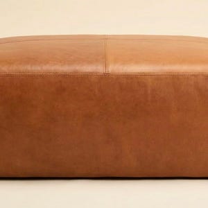 May include: A large, square ottoman upholstered in brown leather. The ottoman has a low profile with rounded corners and a stitched seam detail across the top. The leather has a smooth, slightly textured appearance.