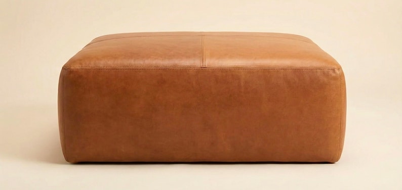 May include: A large, square ottoman upholstered in brown leather. The ottoman has a low profile with rounded corners and a stitched seam detail across the top. The leather has a smooth, slightly textured appearance.