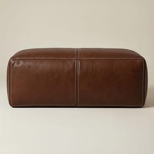 May include: A rectangular brown leather ottoman with white stitching. The ottoman has a padded top and sides, and is designed for use as a footrest or extra seating. The leather has a rich, warm tone.