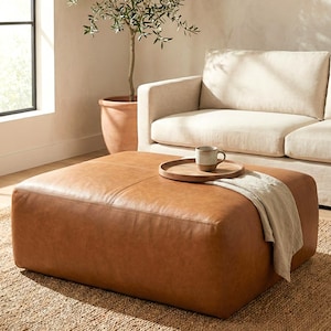 May include: A large, square, brown leather ottoman with stitched detailing. A wooden tray with a mug and a folded blanket rest on top. A beige sofa and a potted olive tree are in the background, creating a cozy living room scene.
