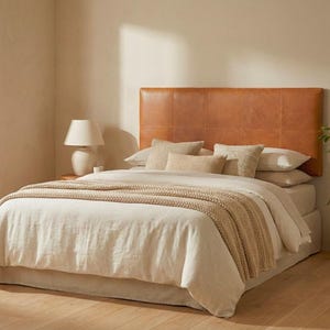 Peut inclure: Un lit avec une tête de lit en cuir marron, une literie crème et une couverture en tricot beige. Le lit est fait avec des oreillers et une lampe est posée sur une table de chevet. Une petite plante en pot est à droite.