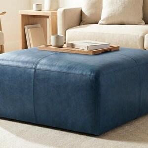 May include: A large, square, blue leather ottoman with stitched detailing. A wooden tray with a book and mug sits on top. The ottoman is in a living room setting with a beige sofa and wooden side table.