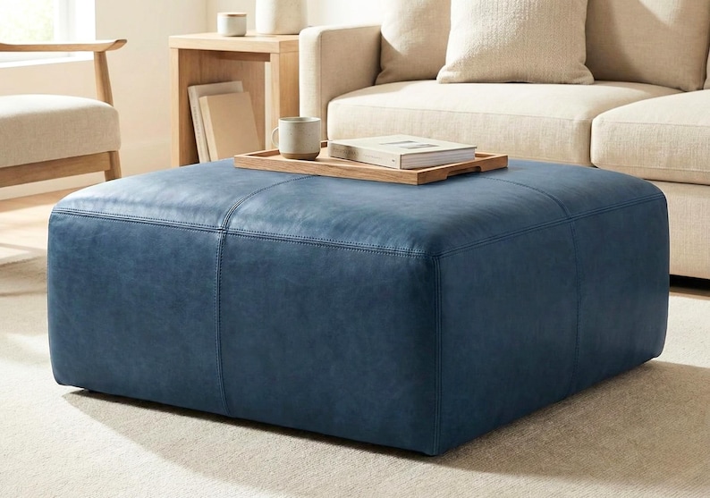 May include: A large, square, blue leather ottoman with stitched detailing. A wooden tray with a book and mug sits on top. The ottoman is in a living room setting with a beige sofa and wooden side table.