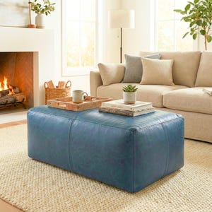 May include: A rectangular, blue leather ottoman sits on a neutral-colored rug in a living room setting. A wooden tray with a mug rests on the ottoman, along with books and a small potted plant. A beige sofa and fireplace are in the background.