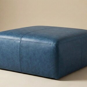 May include: A square, blue leather ottoman. The ottoman has a stitched design and is a solid color. The furniture piece is a low, square shape, and is likely used for seating or as a footrest. The background is a neutral color.