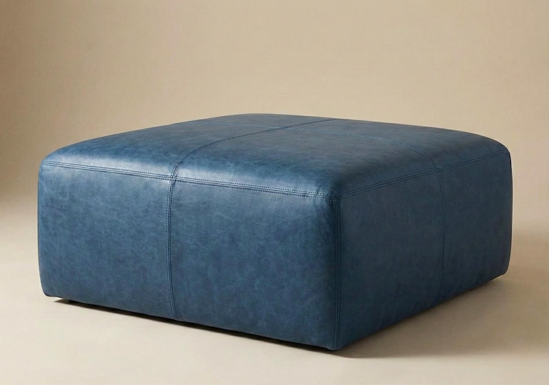 May include: A square, blue leather ottoman. The ottoman has a stitched design and is a solid color. The furniture piece is a low, square shape, and is likely used for seating or as a footrest. The background is a neutral color.