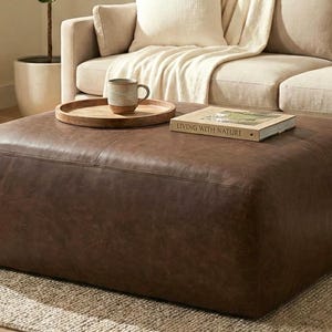May include: A large, square, brown leather ottoman. A wooden tray with a mug sits on top, along with a book titled "Living with Nature." The ottoman is in front of a beige sofa, creating a cozy living room setting.