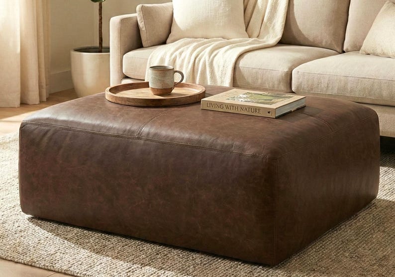 May include: A large, square, brown leather ottoman. A wooden tray with a mug sits on top, along with a book titled "Living with Nature." The ottoman is in front of a beige sofa, creating a cozy living room setting.