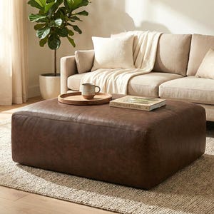 May include: A large, square, brown leather ottoman sits on a neutral-toned rug in front of a beige sofa. A wooden tray with a mug and a book titled "LIVING WITH NATURE" rest on the ottoman. A potted plant is visible in the background.