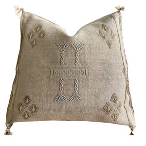 May include: Beige square decorative pillow with embroidered geometric patterns and tassels on each corner. The pillow features diamond and vertical line designs in shades of beige, gold, and blue. The pillow is made of a textured fabric.