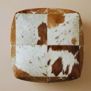 Könnte beinhalten: Quadratischer Hocker aus Kuhfell mit Patchwork-Design. Der Hocker hat eine Mischung aus braunen und weißen Flecken, die mit sichtbaren Nähten zusammengenäht sind. Das Kuhfell hat ein natürliches, strukturiertes Aussehen.