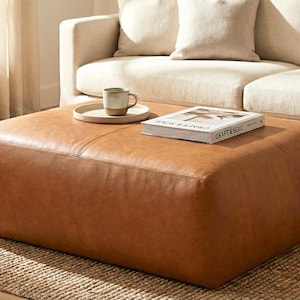 May include: A large, square, brown leather ottoman with a book and a mug on a tray. The ottoman is placed in front of a beige sofa. The book on the ottoman is titled "Craft & Soul". The ottoman is on a woven rug.