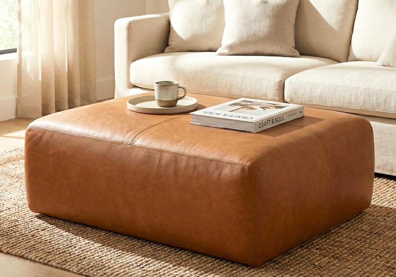 May include: A large, square, brown leather ottoman with a book and a mug on a tray. The ottoman is placed in front of a beige sofa. The book on the ottoman is titled "Craft & Soul". The ottoman is on a woven rug.