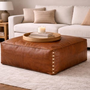 Può includere: Un grande pouf quadrato in pelle marrone con cuciture decorative sul lato. Un vassoio di legno con libri e una candela accesa è posto sopra. Il pouf è davanti a un divano bianco con cuscini beige.